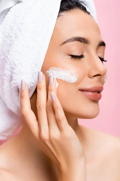 Beautiful woman with towel on hair, closed eyes and cosmetic cream on face isolated on pink — Stock Photo