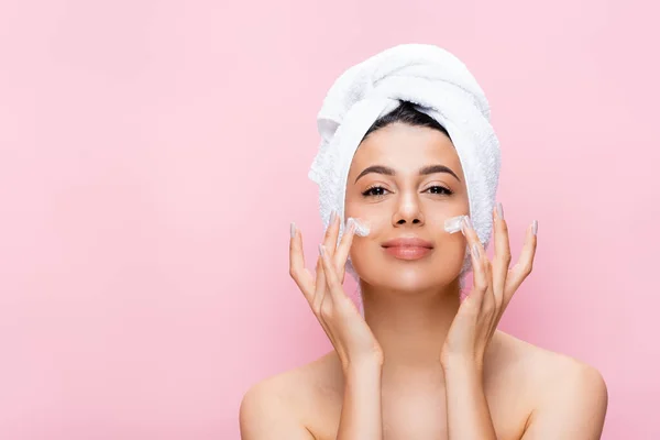 Schöne Frau mit Handtuch auf dem Haar und kosmetische Creme im Gesicht isoliert auf rosa — Stockfoto