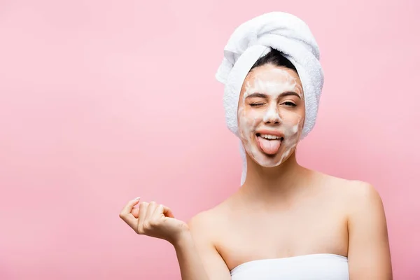 Belle femme avec serviette sur cheveux et mousse sur le visage montrant la langue isolée sur rose — Photo de stock