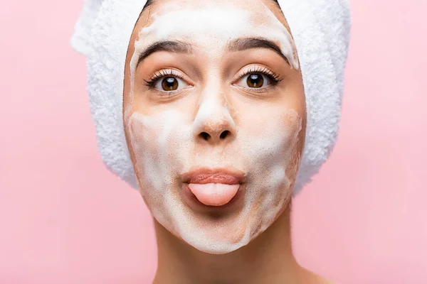 Belle femme avec serviette sur cheveux et mousse sur le visage montrant la langue isolée sur rose — Photo de stock