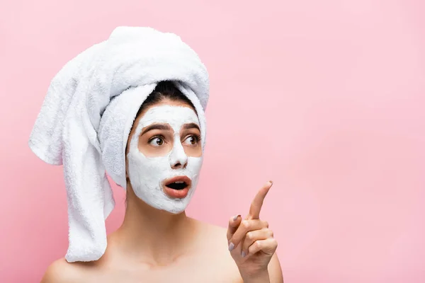 Shocked beautiful woman with towel on head and clay mask on face pointing aside isolated on pink — Stock Photo
