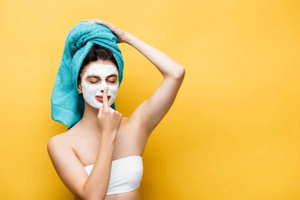 Bella donna con asciugamano blu sui capelli e maschera di argilla sul viso toccare il naso isolato sul giallo — Foto stock