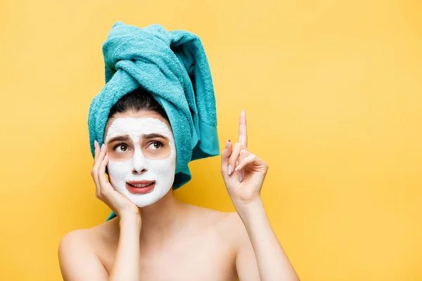 Verwirrte schöne Frau mit blauem Handtuch auf dem Haar und Tonmaske im Gesicht, die isoliert auf gelb zeigt — Stockfoto