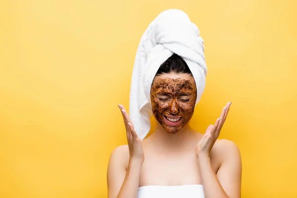 Schöne Frau mit Handtuch auf dem Kopf und Kaffeemaske auf dem Gesicht isoliert auf gelb — Stockfoto