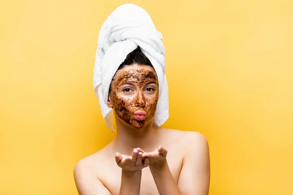 Beautiful woman with towel on head and coffee mask on face blowing kiss isolated on yellow — Stock Photo