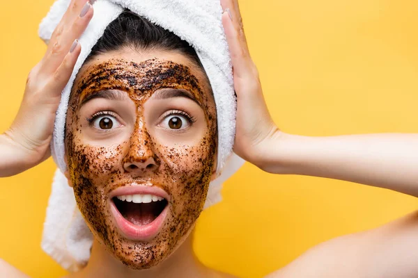 Bella donna sorpresa con asciugamano sulla testa e maschera di caffè sul viso isolato sul giallo — Foto stock