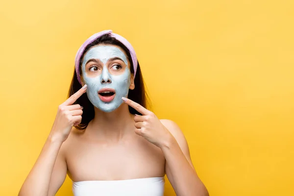 Impactado hermosa mujer apuntando a la máscara de arcilla en la cara aislada en amarillo - foto de stock