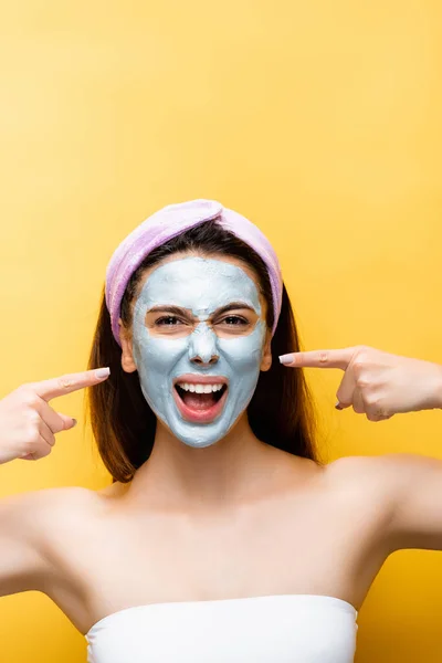 Beautiful woman pointing at clay mask on face isolated on yellow — Stock Photo