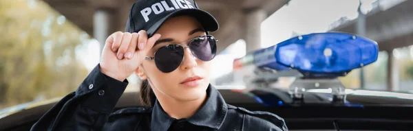 Retrato de una joven policía confiada mirando hacia otro lado cerca del coche patrulla sobre fondo borroso al aire libre, pancarta - foto de stock