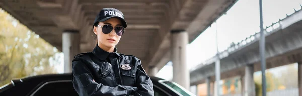 Confident young policewoman with crossed arms looking at camera on blurred background on urban street, banner — Stock Photo