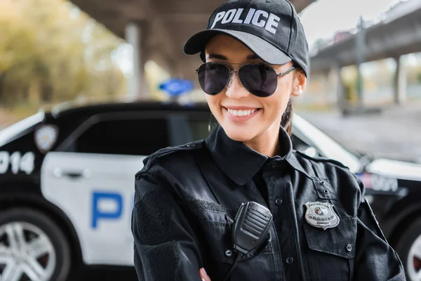 Jovem policial feliz olhando para a câmera com carro patrulha borrada no fundo ao ar livre — Fotografia de Stock