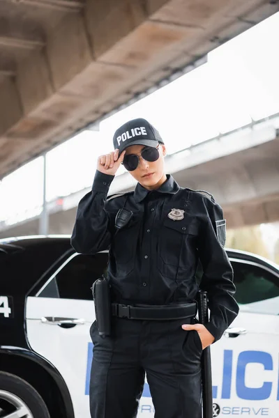 Jeune policière avec la main dans la poche regardant la caméra près de la voiture de patrouille sur fond flou à l'extérieur — Photo de stock
