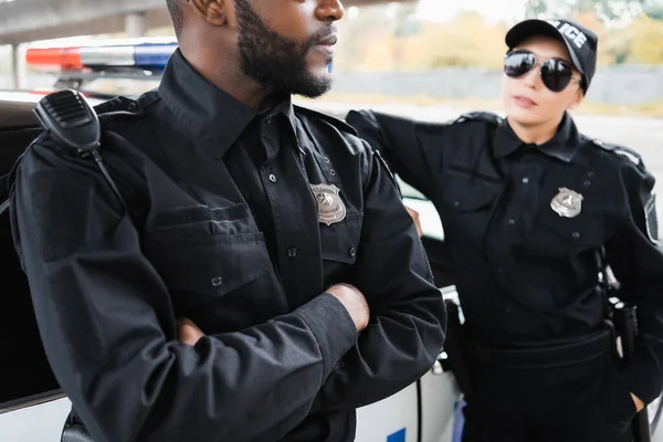 Joven mujer policía mirando a un colega afroamericano con brazos cruzados cerca de patrulla sobre fondo borroso - foto de stock