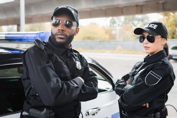 Confiados agentes de policía multiculturales con los brazos cruzados mirando hacia otro lado cerca de la patrulla de automóviles en el fondo borroso al aire libre - foto de stock