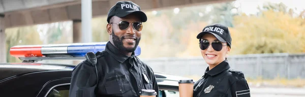 Oficiales de policía multiculturales felices con vasos de papel mirando a la cámara cerca de patrulla sobre fondo borroso al aire libre, pancarta - foto de stock