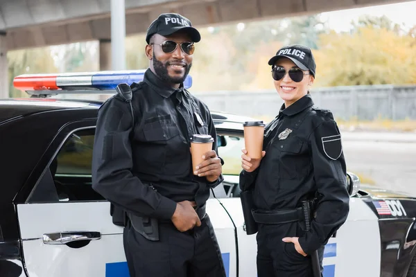 Oficiales de policía multiculturales felices con vasos de papel mirando a la cámara cerca de patrulla sobre fondo borroso en la calle urbana - foto de stock