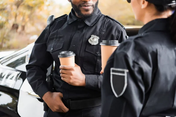 Cropped view of african american policeman with paper cup leaning on patrol car near colleague on blurred background — Stock Photo