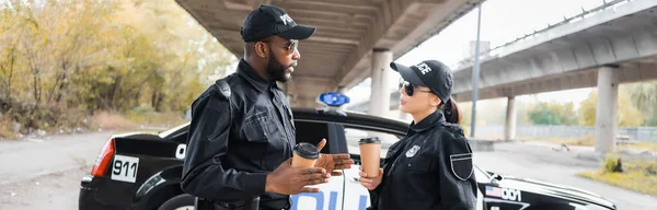 Policías multiculturales con vasos de papel hablando cerca de patrulla sobre fondo borroso en la calle urbana, pancarta - foto de stock