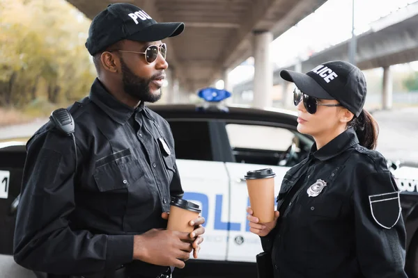 Multicultural police officers with paper cups talking with blurred patrol car on background outdoors — Stock Photo