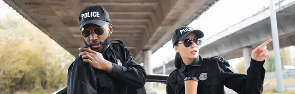 Policewoman apontando com o dedo perto de colega afro-americano falando no rádio set no fundo borrado ao ar livre, banner — Fotografia de Stock