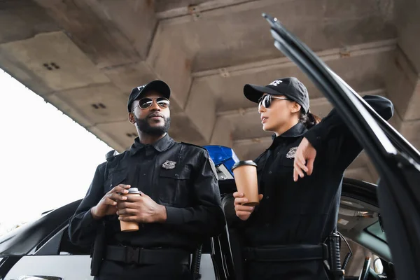 Low angle view of multicultural police officers with paper cups leaning on patrol car on blurred background outdoors — Stock Photo