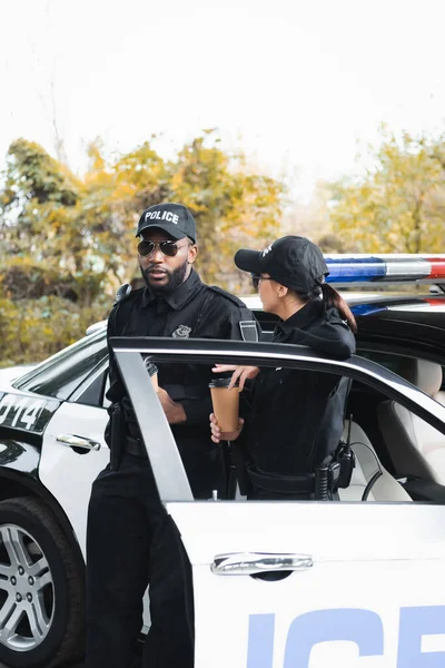 Multicultural police officers with paper cups talking while leaning on patrol car on blurred background outdoors — Stock Photo