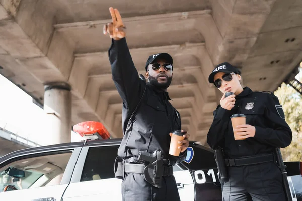 African american policeman pointing with fingers near colleague talking on radio set near patrol car on blurred background — Stock Photo