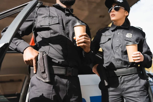 Multicultural police officers with hands on guns, holding paper cups near patrol car on blurred background outdoors — Stock Photo