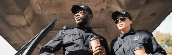 Vista basso angolo di agenti di polizia multiculturali con tazze di carta guardando altrove su sfondo sfocato all'aperto, banner — Foto stock