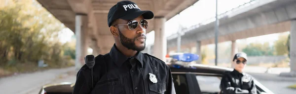 Серьезный африканский американский полицейский, глядя в сторону с размытым коллегой на заднем плане на городской улице, баннер — стоковое фото