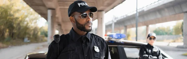 Sério policial afro-americano olhando para longe com colega turvo no fundo na rua urbana, banner — Fotografia de Stock