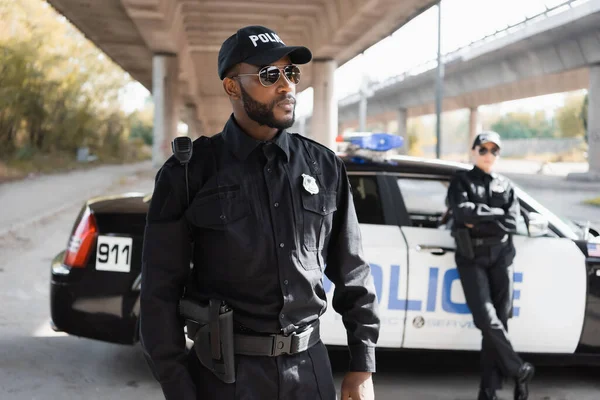 Серйозний афро-американський полісмен, який дивиться з розмитим колегою і патрульною машиною на вулиці міста — стокове фото