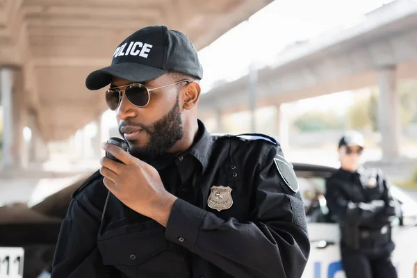 Афроамериканський поліцейський говорить по радіо, встановленому біля поліцейської на розмитому фоні на відкритому повітрі — стокове фото