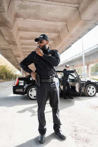 In voller Länge afrikanisch-amerikanischer Polizist mit der Hand an der Waffe im Gespräch im Radio neben Kollegen und Streifenwagen auf verschwommenem Hintergrund im Freien — Stockfoto