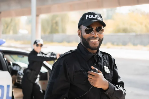 Happy african american police officer looking at camera while holding radio set on blurred background outdoors — Stock Photo