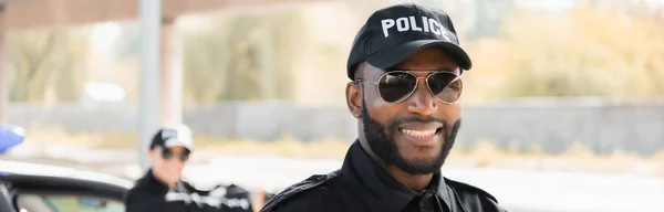 Porträt eines glücklichen afrikanisch-amerikanischen Polizisten, der in die Kamera blickt, mit verschwommenem Kollegen im Hintergrund im Freien, Banner — Stockfoto