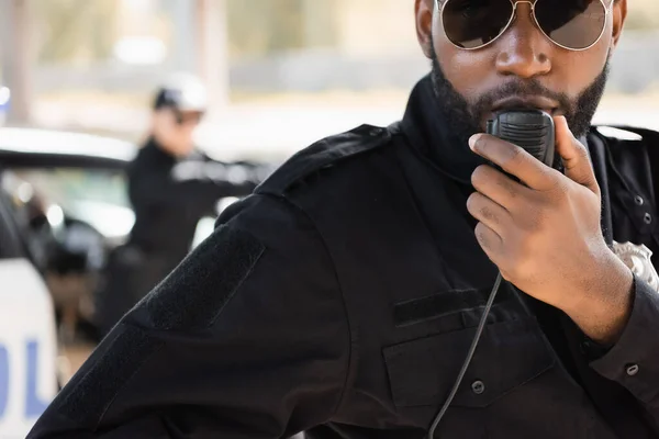 Nahaufnahme eines afrikanisch-amerikanischen Polizisten, der am Funkgerät mit einem verschwommenen Kollegen im Hintergrund im Freien spricht — Stockfoto