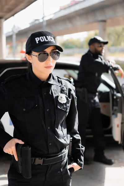 Confiada mujer policía con la mano en el arma mirando hacia otro lado con borrosa colega afroamericano en el fondo al aire libre - foto de stock