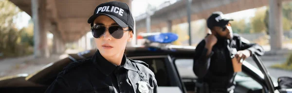 Serious policewoman looking away with blurred african american colleague on background outdoors, banner — Stock Photo