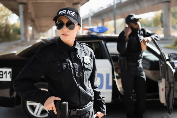 Серьезная женщина-полицейский с пистолетом, глядя в сторону африканского американского коллеги на размытом фоне на улице — стоковое фото