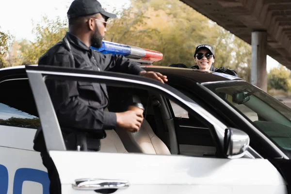 Happy policewoman leaning on patrol car near african american colleague on blurred background outdoors — Stock Photo