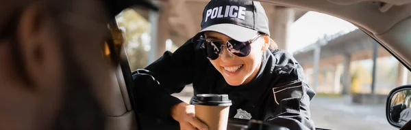 Happy policewoman with paper car looking at african american colleague in patrol car on blurred foreground, banner — Stock Photo