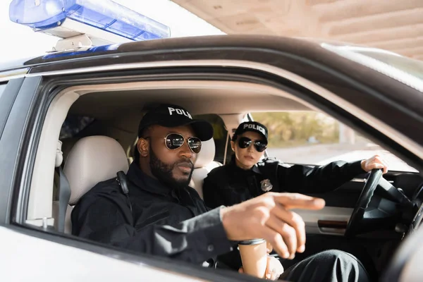 Policier afro-américain pointant du doigt un collègue proche dans une voiture de patrouille sur fond flou — Photo de stock