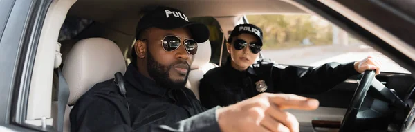 African american policeman pointing with finger near colleague in patrol car on blurred background, banner — Stock Photo