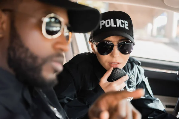 Policier parler sur le poste de radio avec un collègue afro-américain flou au premier plan en voiture de patrouille — Photo de stock