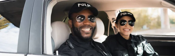 Happy multicultural police officers looking at camera in patrol car on blurred background, banner — Stock Photo