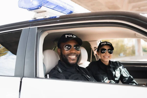 Sorrindo policiais multiculturais olhando para a câmera em fundo embaçado em carro de patrulha — Fotografia de Stock