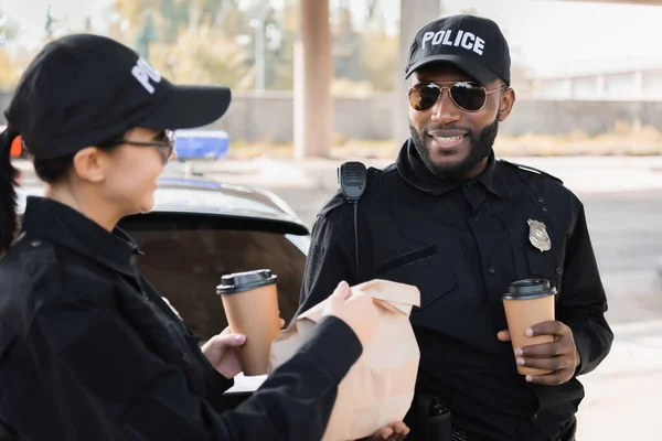 Felice poliziotta con coppa di carta dando pacchetto al collega africano americano vicino pattuglia auto su sfondo sfocato all'aperto — Foto stock