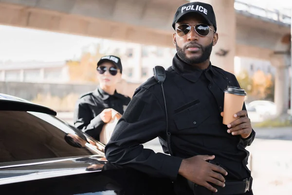 Policier afro-américain avec la main sur l'arme regardant loin avec un collègue flou en arrière-plan à l'extérieur — Photo de stock