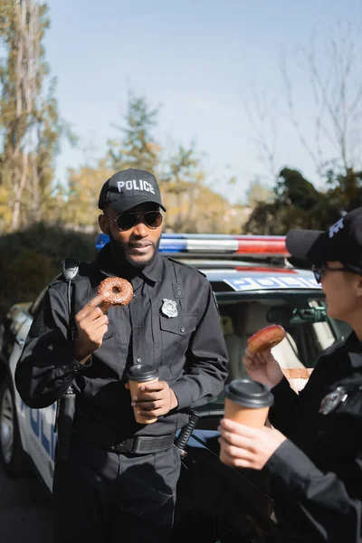 African american police officer with doughnut and paper cup looking at colleague near patrol car on blurred background outdoors — Stock Photo